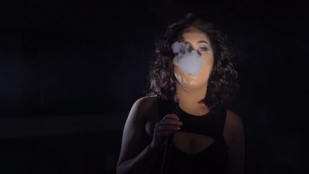 Retrato de glamour horizontal. A mulher de cabelo preto é arrepiante fumando o narguilé e produzindo a fumaça grossa no estúdio preto . — Vídeo de Stock