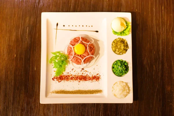 Mâncare gustoasă din carne crudă, gălbenuș de ou, verdeață și condimente plasate pe placa dreptunghiulară albă. Viziunea de mai sus . — Fotografie, imagine de stoc