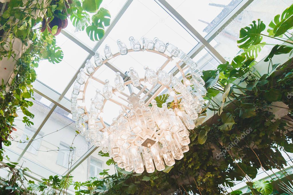 The upside close-up photo of the huge modern chandelier made of the empty bottles surrounded by green plants.