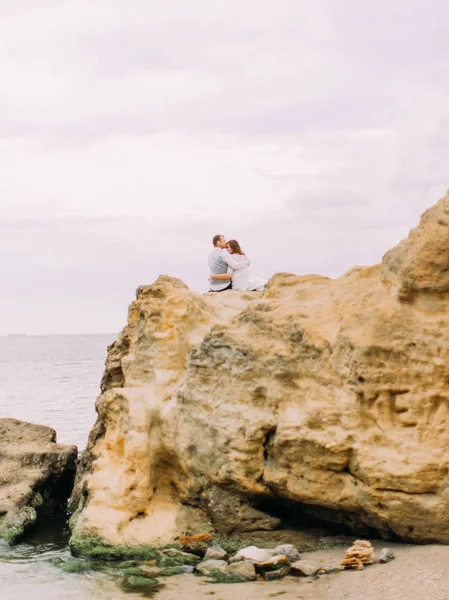 La vista verticale degli sposi seduti sulle rocce tra il mare. Lo sposo sta baciando la sposa sulla testa . — Foto Stock