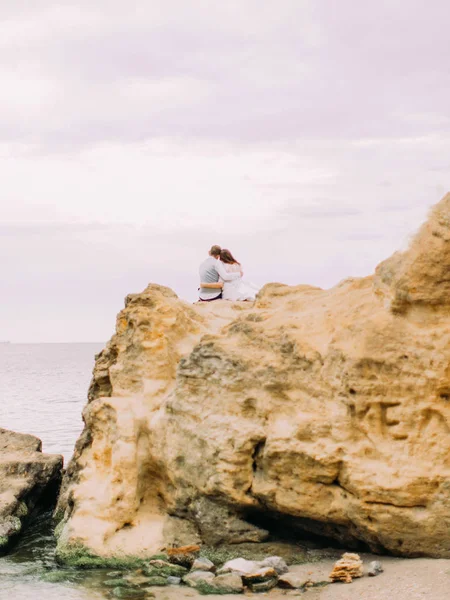 La parte posteriore degli sposi che si abbracciano mentre siedono sulla scogliera e guardano il tramonto nel mare . — Foto Stock