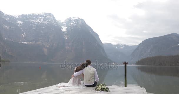 Rückansicht des schönen küssenden Paares, das auf der Seebrücke sitzt und die Aussicht auf die Berge genießt. — Stockvideo