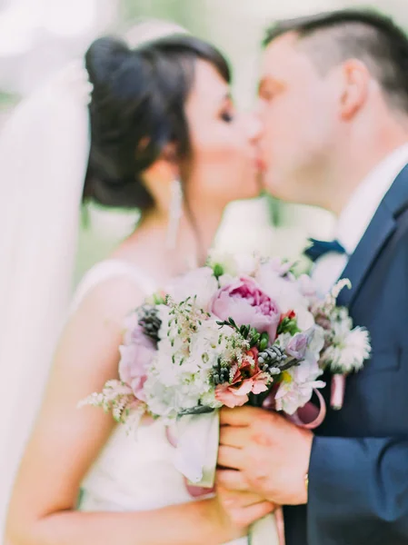 Schöner Hochzeitsstrauß aus farbenfrohen Pfingstrosen mit Kräutern in den Händen küssender Brautpaare. — Stockfoto