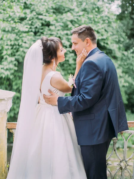 La vue arrière du couple de jeunes mariés heureux. La mariée caresse la joue du marié dans le jardin . — Photo