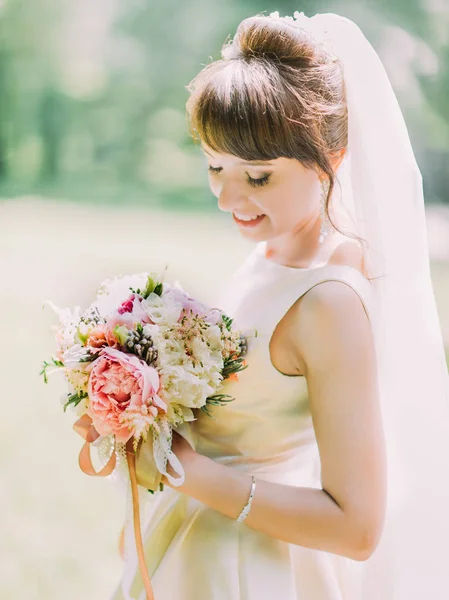 Retrato de cerca de la novia sonriente mirando hacia abajo y sosteniendo el ramo de bodas de coloridas peonías. Ubicación del parque . — Foto de Stock