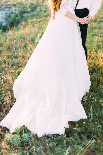 A parte de baixo do vestido de casamento longo da noiva e o noivo que está na grama verde coberta de flores . — Fotografia de Stock