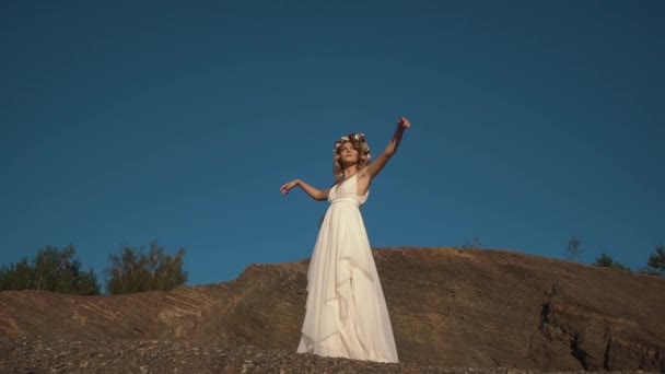 Красива дівчина в довгій білій сукні в стилі бохо і з вінком квітів на голові проводить час в горах — стокове відео