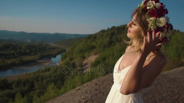 Die hübsche Frau im weißen Boho-Kleid vor dem Hintergrund der herrlichen Berge, berührt den Blumenkranz auf ihrem Kopf — Stockvideo