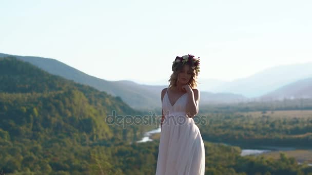 Приваблива блондинка з квітковим віночком і в білій сукні бохо задоволена сонячним днем у горах — стокове відео