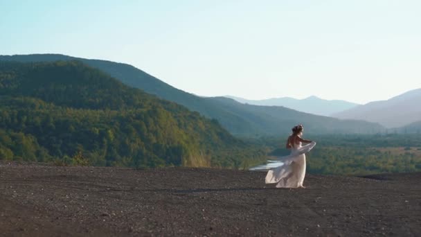 La mujer en el vestido largo boho blanco y con la corona de flores en la cabeza está caminando al fondo de las montañas y el río — Vídeos de Stock