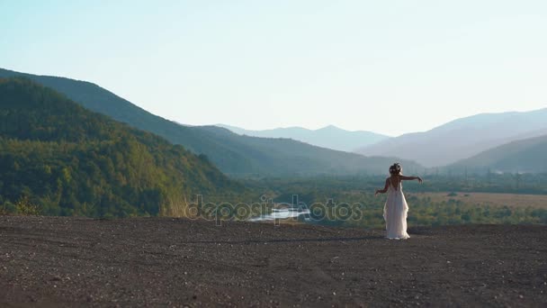 白色长波西米亚礼服的年轻妇女的放松和她的头上的花花圈在绿山 — 图库视频影像