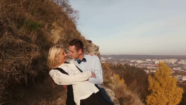 Мужчина целует девушку, сидящую на скале перед городом — стоковое видео