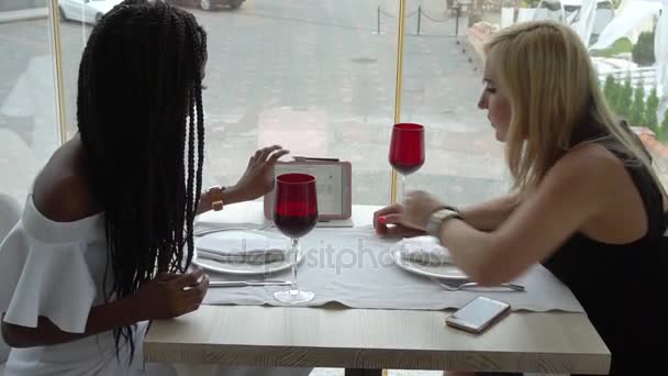 Две женщины пьют вино из красных бокалов на неформальной встрече в ресторане, печатая на планшете и разговаривая — стоковое видео
