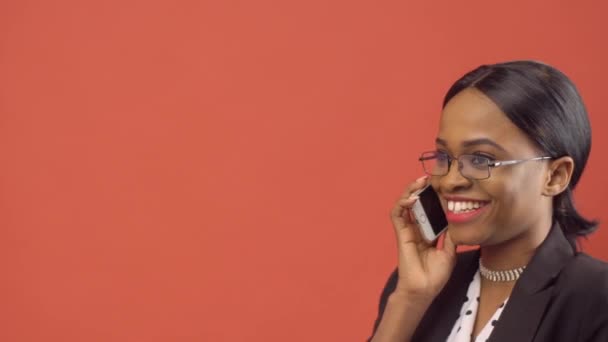 Mulher negra em óculos fala em seu iPhone sentado diante da parede vermelha — Vídeo de Stock