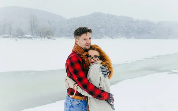 Retrato sensível do homem bonito abraçando seu amante da cabeça vermelha no prado coberto de neve durante a queda de neve. Tempo de Natal . — Fotografia de Stock