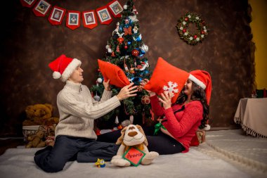 Noel Baba şapkaları mutlu kapalı aile portresi. Güzel hamile kadın kocasıyla dekore edilmiş Noel ağacı yakınındaki yastıklar oynuyor.