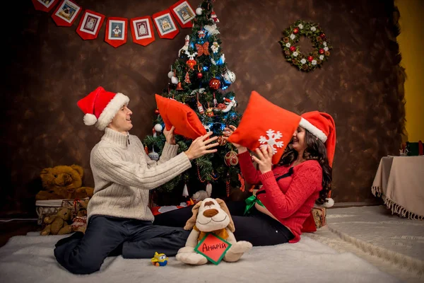 快乐的室内家庭人像圣诞老人的帽子。美丽的孕妇正在与她的丈夫在装饰圣诞树附近玩枕头. — 图库照片