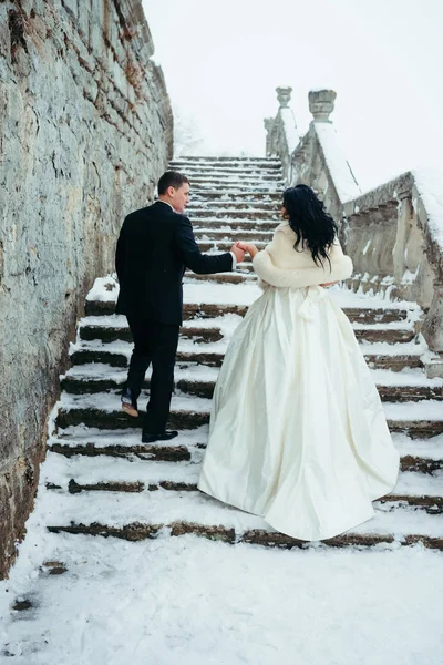 The full-length back view of the attractive newlywed couple holding hands and going up the old stairs covered with fluffy snow.