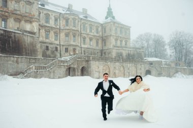 Mutlu gülen gelin damadın elini tutuyor ve onu eski kalenin arka plan, karlı çayır boyunca çalıştırırken lider.