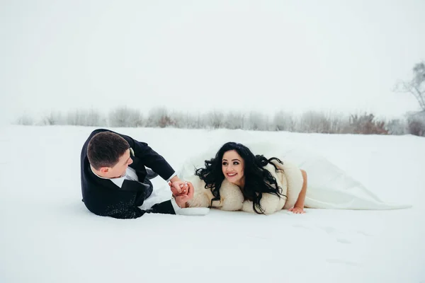 Divertido retrato al aire libre de los felices recién casados sonrientes que yacen en la nieve . — Foto de Stock