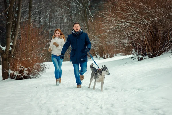 Alegre pareja amorosa con husky siberiano está corriendo a lo largo del bosque nevado durante su paseo . — Foto de Stock