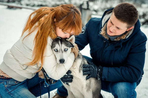 Sensible retrato de cerca de la mujer pelirroja besándose encantadora husky siberiana mientras el hombre lo acaricia. Tiempo de invierno . — Foto de Stock