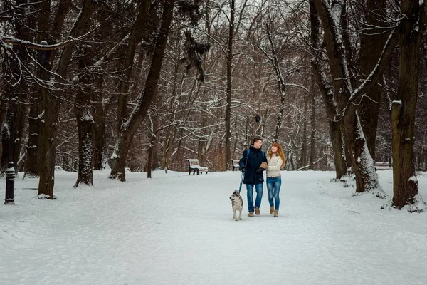 O passeio do casal amoroso feliz com seu cão siberiano ao longo do caminho nevado. Tiro horizontal . — Fotografia de Stock