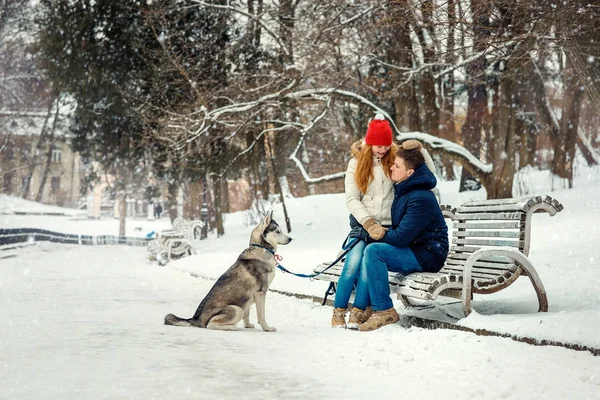 La pareja feliz está pasando su tiempo con el perro siberiano en el parque nevado durante la nevada. La encantadora pelirroja está sentada en las vueltas de su novio . — Foto de Stock