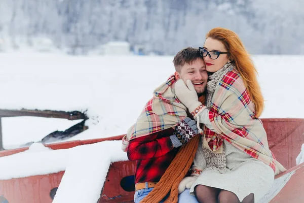 Sensitive half-length portrait of the beautiful young woman stroking the cheek of the handsome man. They are wrapped in the knitted plaid while sitting on the boat during the snowfall. — Stock Photo, Image