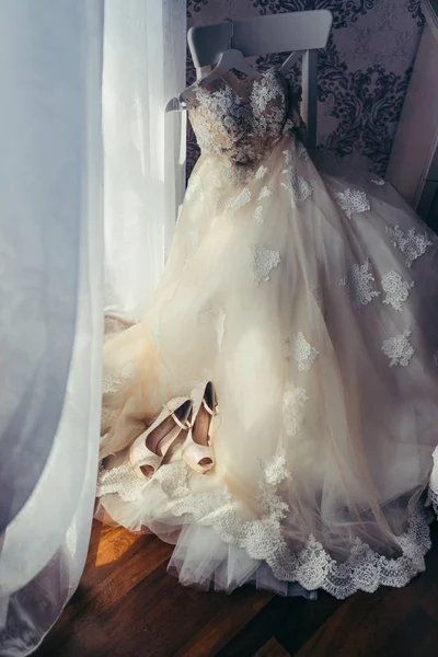 Die vertikale Ansicht des stylischen weißen langen Brautkleides mit Blumen, die auf dem Stuhl hängen. die beigen High Heels sind auf dem Kleid platziert. — Stockfoto