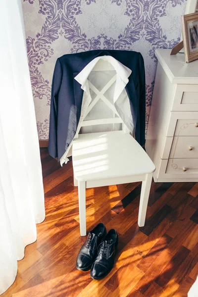 Manhã do noivo. A jaqueta e a camisa do terno estão penduradas na cadeira e os sapatos pretos brilhantes que estão perto dela no quarto ensolarado elegante . — Fotografia de Stock