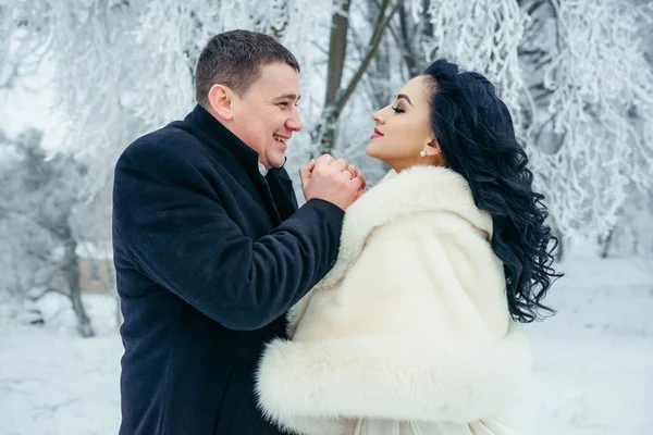 Ritratto sensibile della bella coppia di sposi allegri. Lo sposo sta riscaldando le mani della sua affascinante sposa bruna nella foresta invernale . — Foto Stock