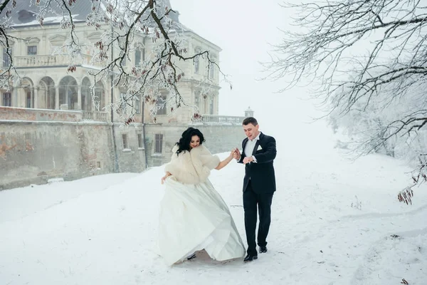 Feliz pareja recién casada está cogida de la mano y caminando alegremente a lo largo del bosque nevado cerca de la antigua casa. Vista de cuerpo entero . — Foto de Stock