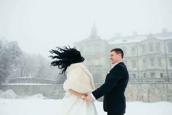 La sposa sta scuotendo i capelli mentre si tiene per mano con lo sposo sorridente nel giorno delle nozze invernali sul prato vicino alla vecchia casa . — Foto Stock