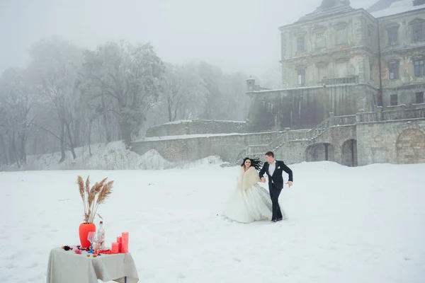 Casal recém-casado alegre está de mãos dadas e correndo alegremente para a mesa organizada para o jantar romântico ao ar livre. Hora de Inverno . — Fotografia de Stock