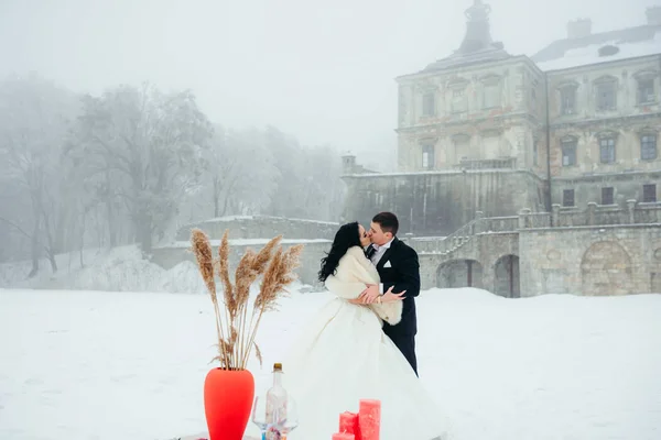 Hermosos recién casados felices se besan cerca de la mesa arreglada para la cena al aire libre romántica en el prado nevado en el fondo de la antigua casa . — Foto de Stock