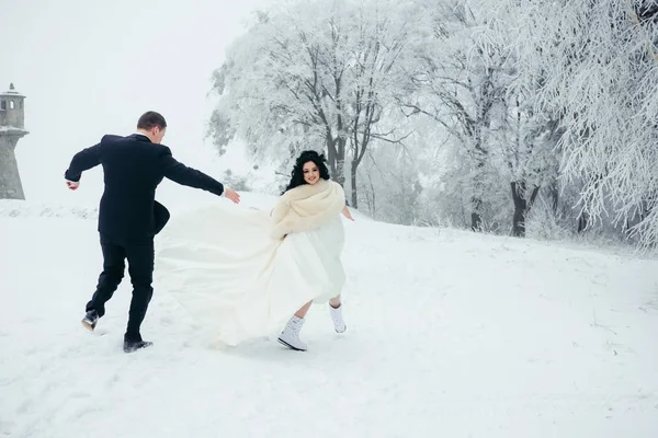 Happy newlywed couple is spending great time in the snowy forest. The groom is running after his beautiful brunette bride. — Stock Photo, Image