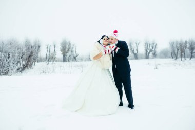 Komik şapkalar ve bardak tutan ve birbirine yakın karlı çayır üzerinde duran eldiven çekici mutlu yeni evliler tam uzunlukta atış.