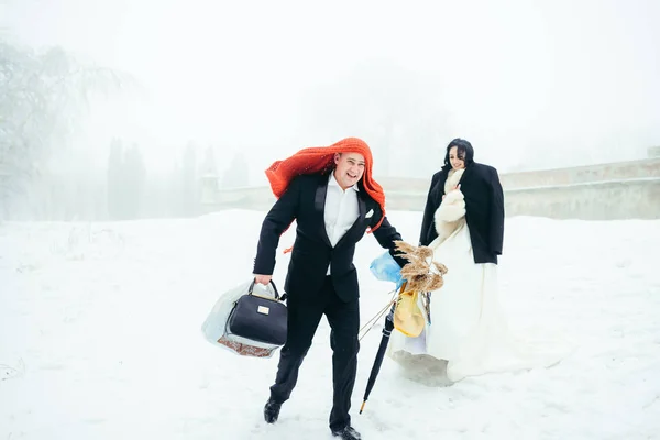Fulle-lenght view of the happy groom with the knitted scarf on the head holding all things for the winter picnic. The beautiful bride is standing behind. — Stock Photo, Image
