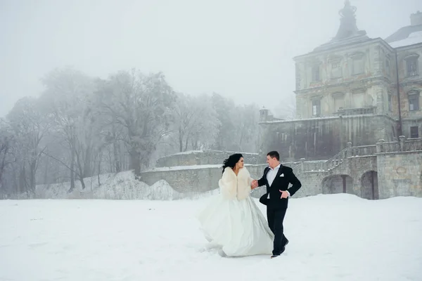 Walking happy newlywed couple along the snowy meadow at the background of the old house. — Stock Photo, Image