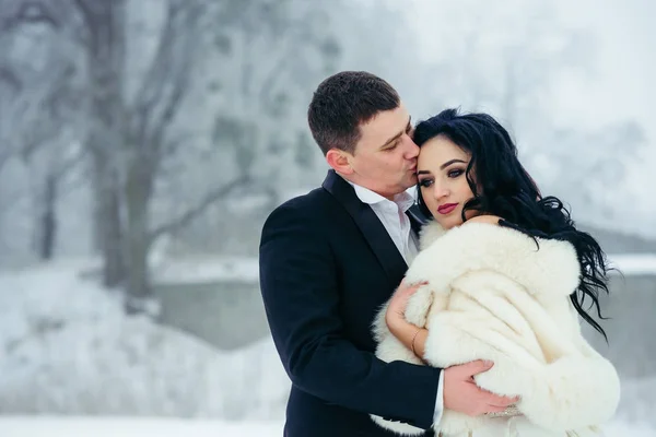 Bruidegom is zacht hugp ouging en zijn charmante brunette bruid kussen in voorhoofd. Close-up buiten winter portret. — Stockfoto