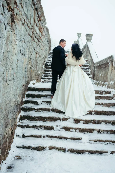 The full-length back view of the happy newlyweds holding hands and going uo the snowy stairs of the ancient house. — Stock Photo, Image