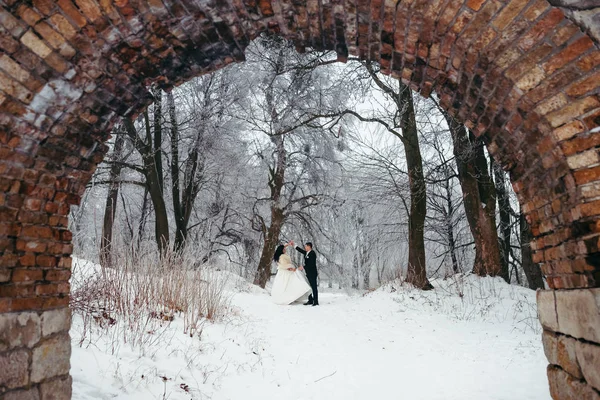 Dancing happy newlywed couple in the snowy forest. View through the old stone arch.