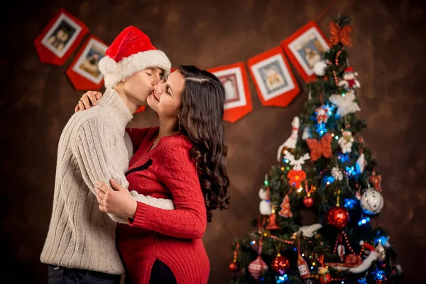 Das halblange Porträt der schönen, fröhlichen zukünftigen Eltern. Der Mann küsst seiner schwangeren Frau im Hintergrund des Weihnachtsbaums auf die Wange. — Stockfoto