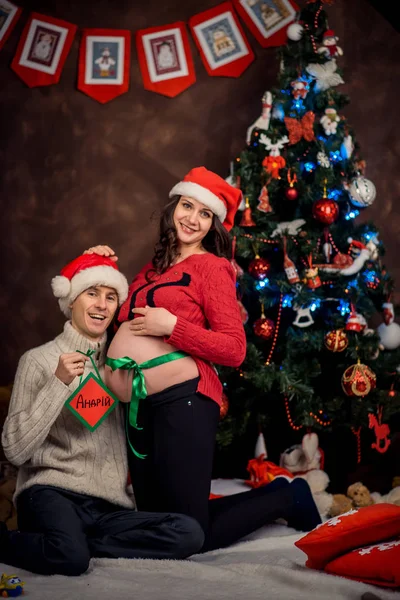 Glückliches Paar Frau schwanger Bauch eingewickelt Bänder umarmen Weihnachtsbaum Neujahr Glück Weihnachtsmann Hüte halten Zeichen andrii. — Stockfoto