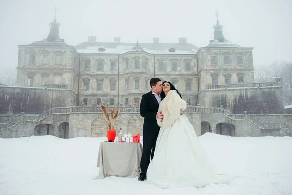 Groom is kissing the beautiful smiling bride in cheek near the arranged table for outdoor dinner on the snowy meadow near the old house. Full-length view. — Stock Photo, Image