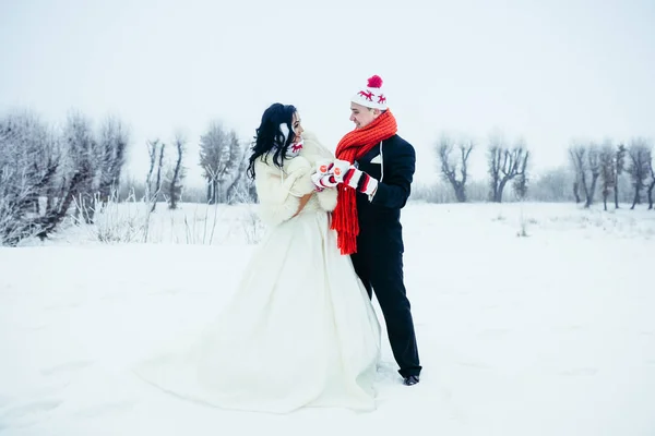 Tiro completo do belo par recém-casado feliz que se diverte no prado nevado. Eles estão usando luvas engraçadas, chapéus e copos de segurando com chá . — Fotografia de Stock