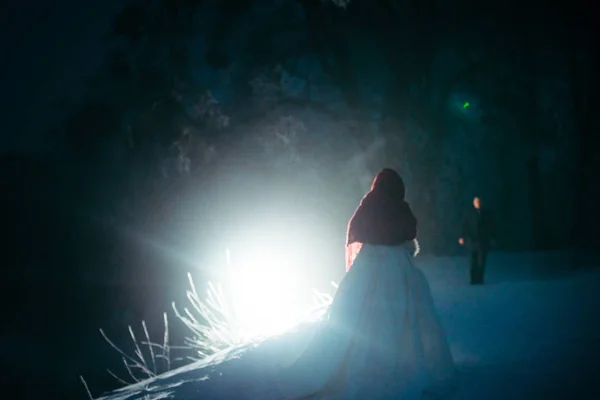 Vista trasera de la novia en bufanda de punto rojo esperando al novio que viene cerca de la luz blanca en el bosque nevado de la noche . — Foto de Stock