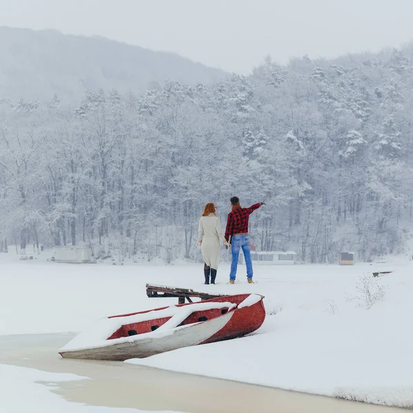 站在船和森林附近的雪地上的手夫妇的背面视图。那个男人正用手向他的爱人展示一些东西. — 图库照片