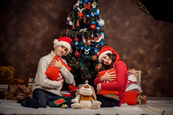 Happy future parents in Santa Claus hats are sitting on the plaid and hugging the pillows at the background of the Christmas tree.